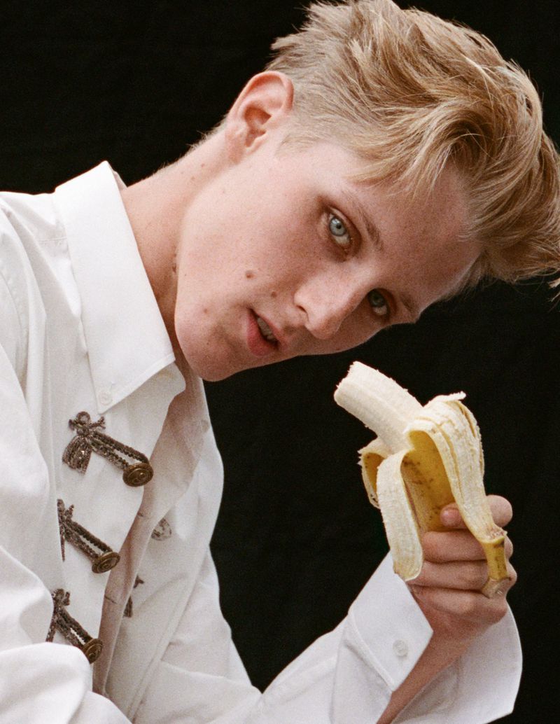 Young adult with short blonde hair, pale skin, holding a partially peeled banana. He is wearing a white collared shirt against a black backdrop.