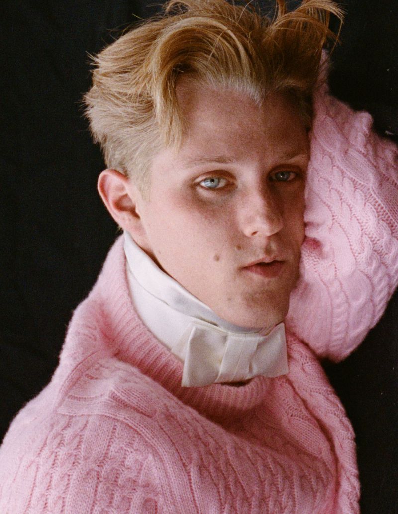 Portrait of a fair-skinned young adult with blond hair wearing a pink knit sweater and a white dress shirt with a bow tie against black.