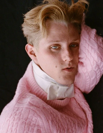 Portrait of a fair-skinned young adult with blond hair wearing a pink knit sweater and a white dress shirt with a bow tie against black.