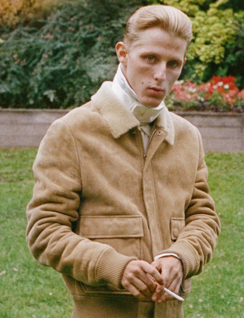 Portrait of a blonde adult man in a tan suede jacket smoking a cigarette outdoors with green foliage and colorful flowers in the background.
