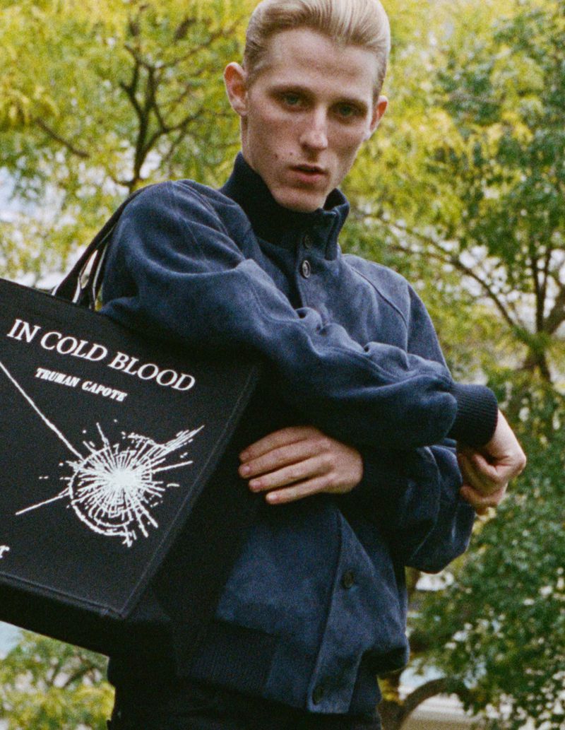 Model Valentin Campagne in a navy blue bomber jacket holding a black printed tote bag outdoors surrounded by green foliage.