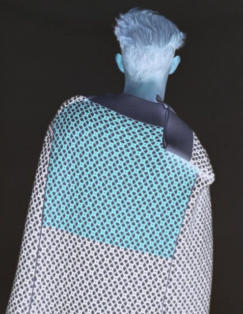 Person from the back wearing patterned cape against dark background. Fashion shot showcasing design and texture. Pattern and garment detail.