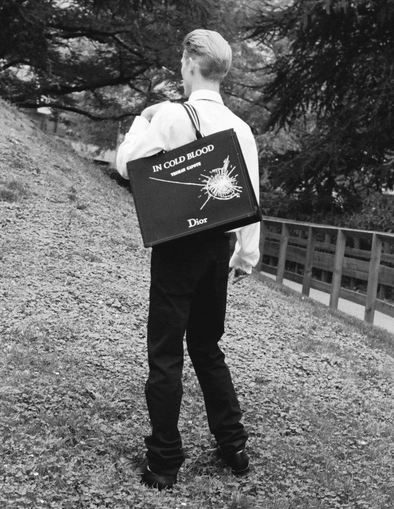Blond-haired person in a white shirt and black pants, walking on a path with a Dior shopping bag on their shoulder.