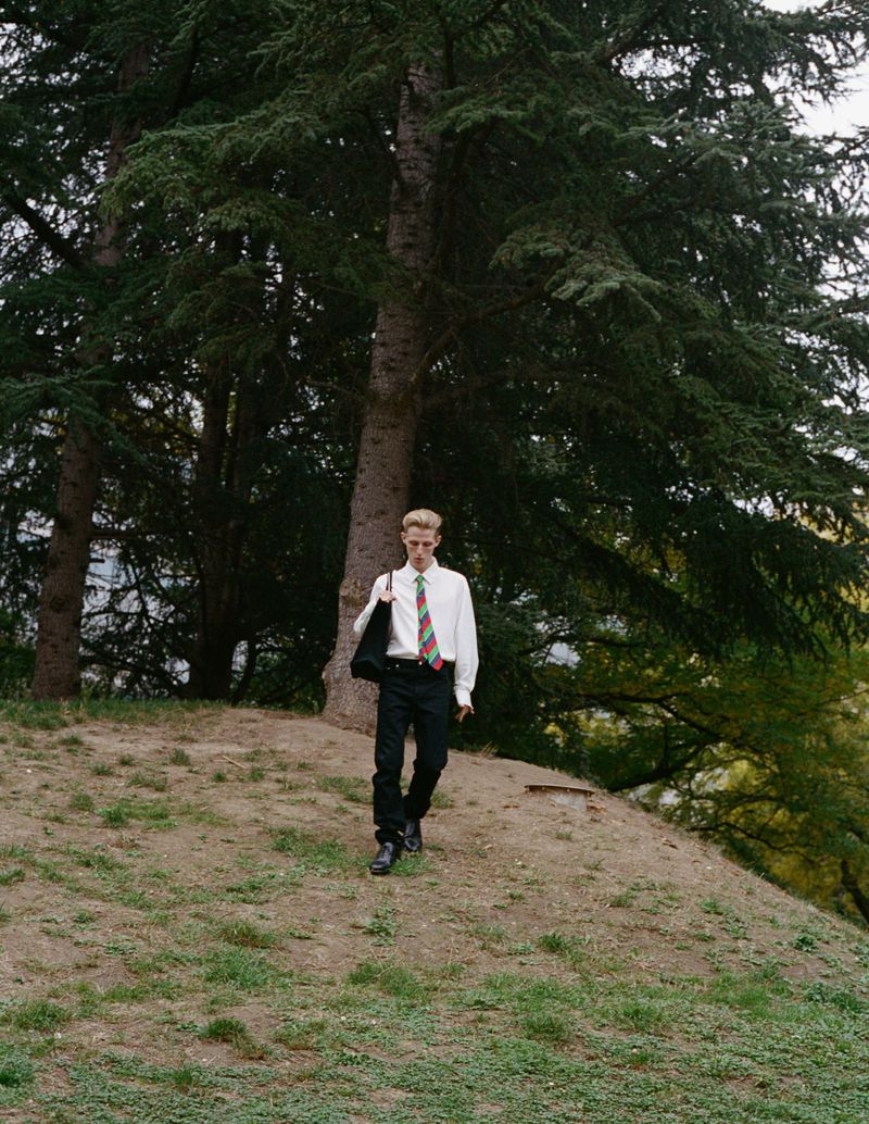 A fair-skinned young adult male walks downhill on a grassy slope near tall trees, wearing black pants, a white shirt, and a patterned tie.