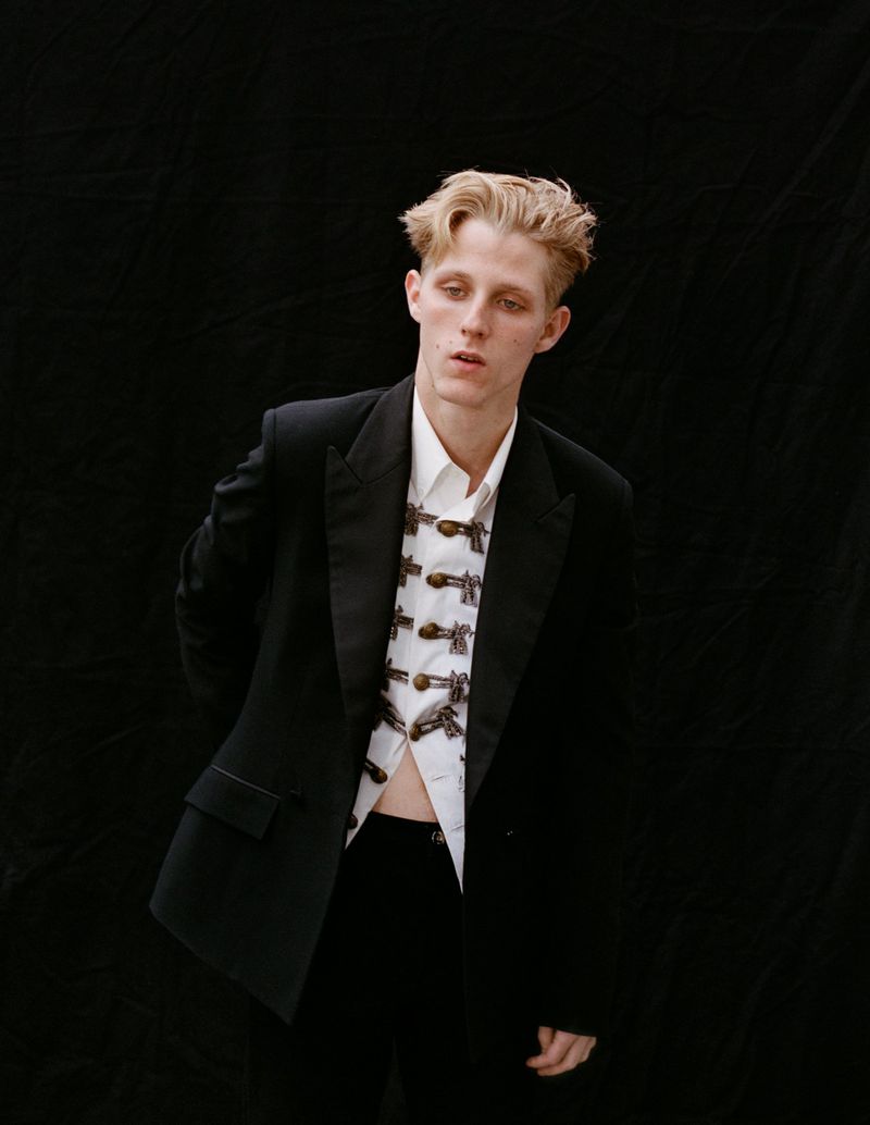 Portrait of a young adult male with blonde hair wearing a black tuxedo jacket and white button-down shirt against a black backdrop.