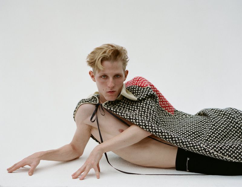 Model lies on white background wearing black shorts and a patterned black, red, and white textile cape with tie strings.