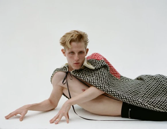 Model lies on white background wearing black shorts and a patterned black, red, and white textile cape with tie strings.