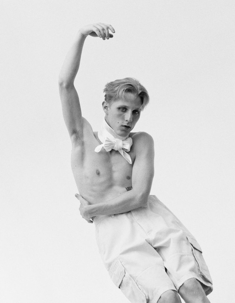 Man in white shorts and bow tie poses shirtless in studio. Black and white fashion portrait with artistic composition, fair skin.