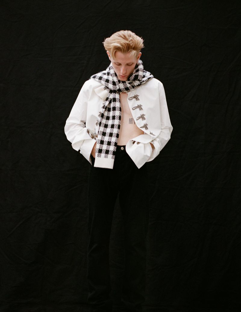 Blond male model wearing a white jacket and black and white scarf posing indoors against a black background, looking down.