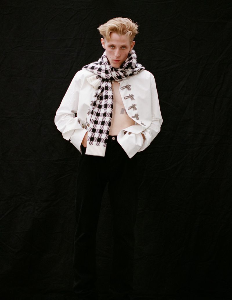 Studio shot of a young adult wearing a white jacket and plaid scarf against a black backdrop, posing in a fashion setting.