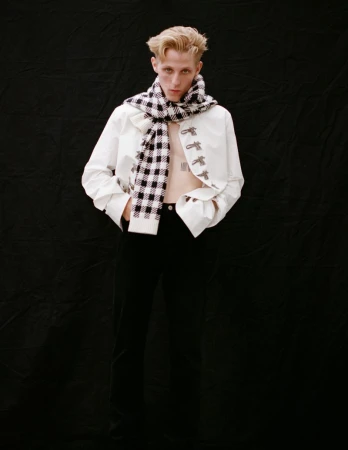 Studio shot of a young adult wearing a white jacket and plaid scarf against a black backdrop, posing in a fashion setting.