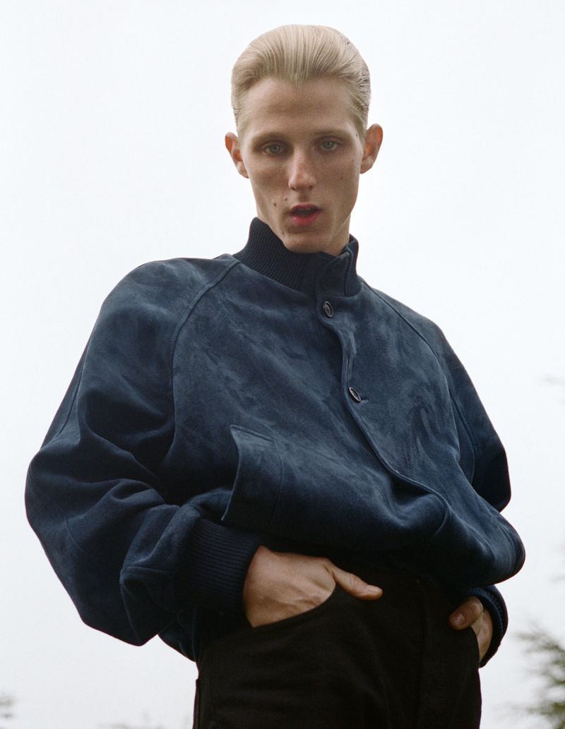 A fair-skinned adult male model with blond hair, wearing a navy bomber jacket and black pants, poses outdoors with a neutral expression.
