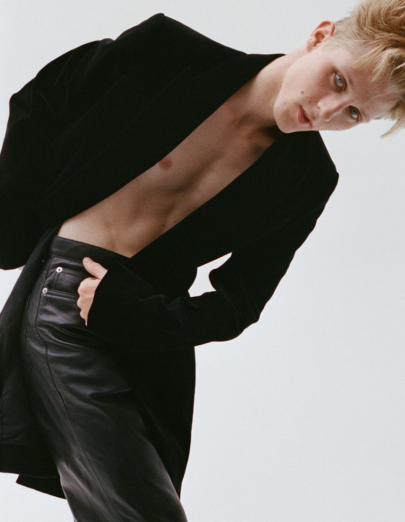 Blonde-haired person in a black blazer and black leather pants leans into frame against a white backdrop in a fashion studio shot.