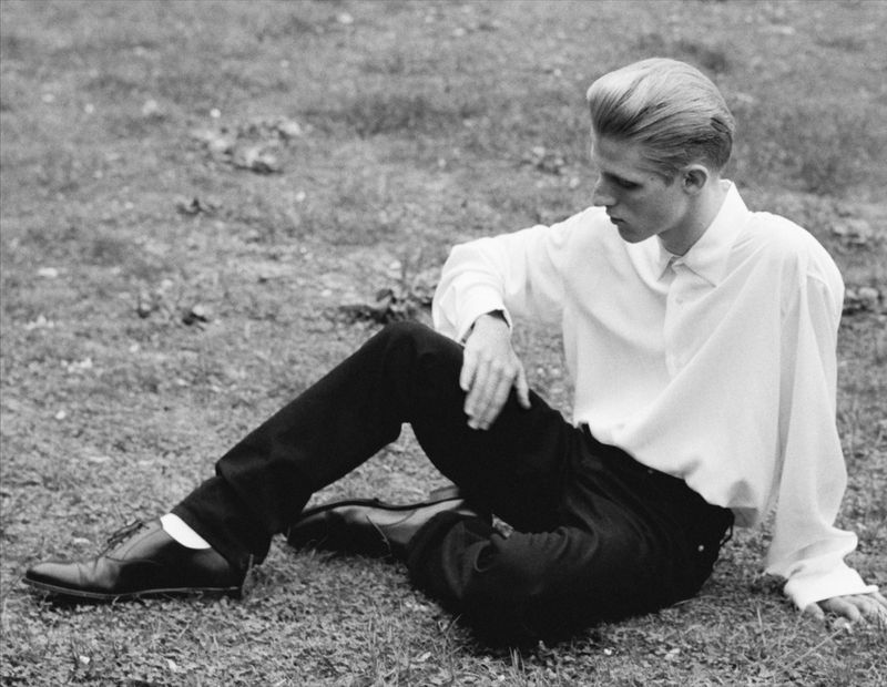 Monochrome outdoor shot of a young adult male in white shirt and dark pants sitting on grass, facing left and looking off to the side.