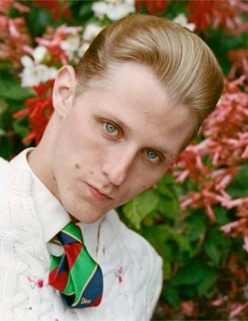Portrait of Valentin Campagne in Dior wearing a white cable-knit sweater and striped tie against red flowers