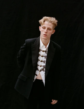 Model Valentin Campagne posing in a black blazer and patterned white shirt against a solid black background.