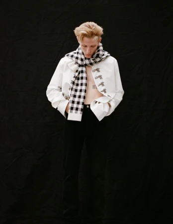 Valentin Campagne in Dior for Arena Homme Plus wearing a white jacket, black trousers, and a black-and-white checked scarf against a black backdrop