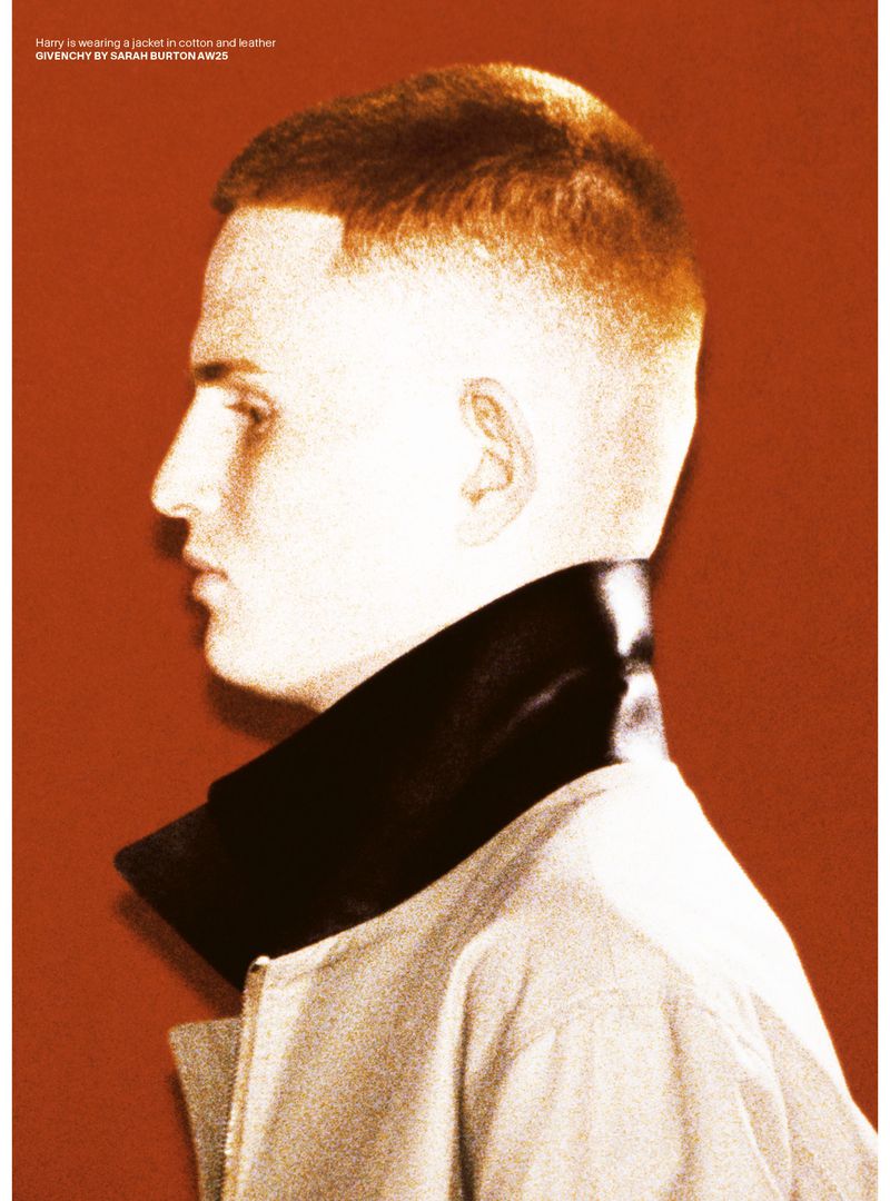 Profile view of an adult male with short hair wearing a jacket with a wide leather collar against a solid orange backdrop.