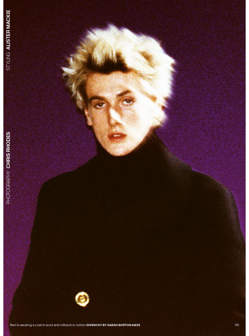 Close-up portrait of a blond male model in black coat and turtleneck against a textured purple background in a fashion editorial.