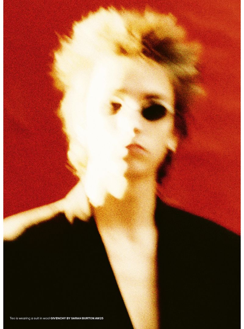 A close-up studio shot shows a person with dark glasses and tousled blonde hair, wearing a dark blazer against a red background.