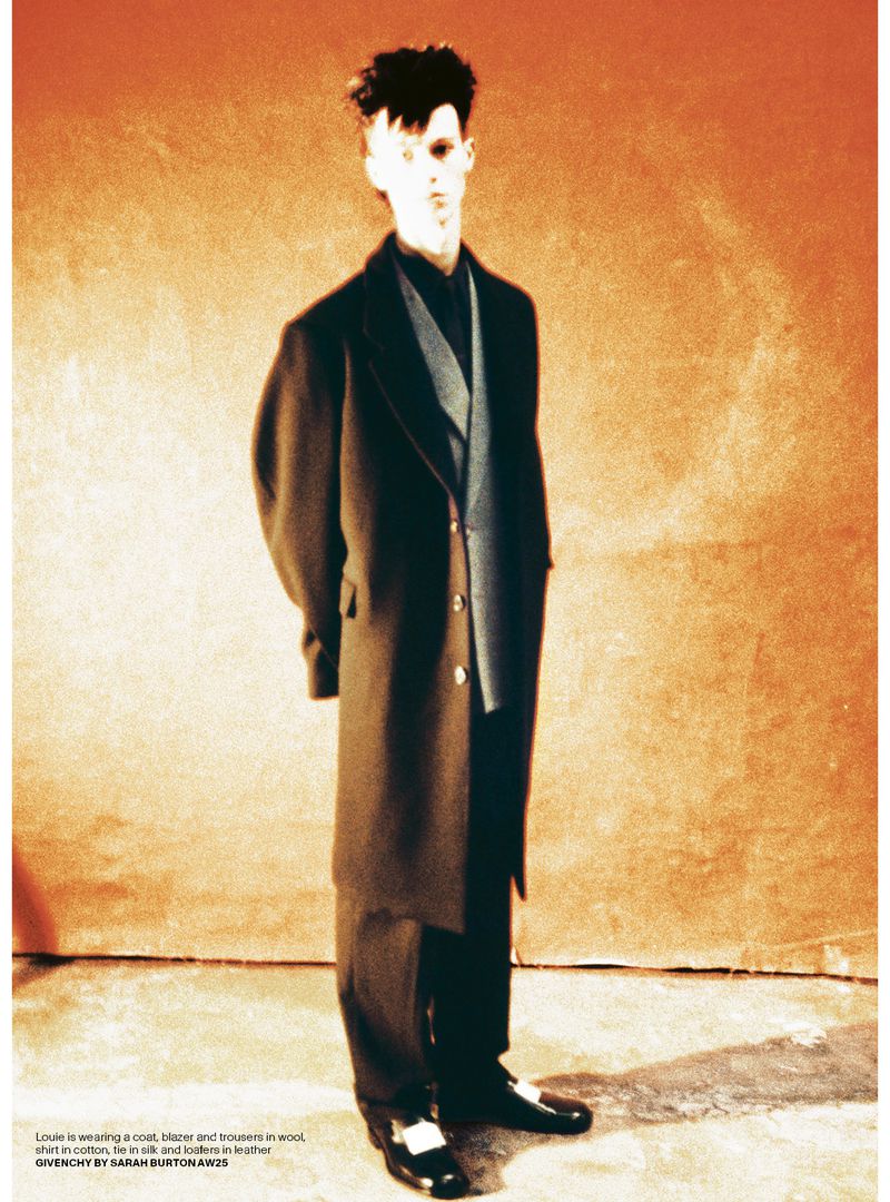 A slender adult male poses in a long dark coat with black pants and shoes against an orange-toned backdrop in a fashion shot.