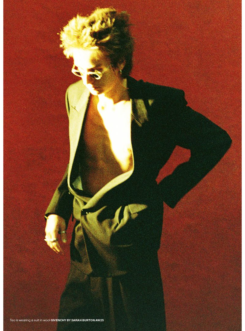 Fashion shot of a young adult with stylish hair wearing a black blazer and skirt against a red backdrop, wearing sunglasses.