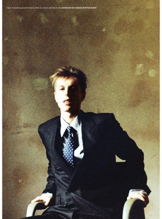 Man in a dark suit and spotted tie sitting in a chair against a textured mottled wall.