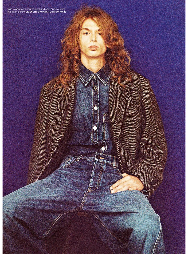 A long-haired male fashion model poses while wearing a denim shirt and jeans with a wool coat, in a portrait against a violet backdrop.