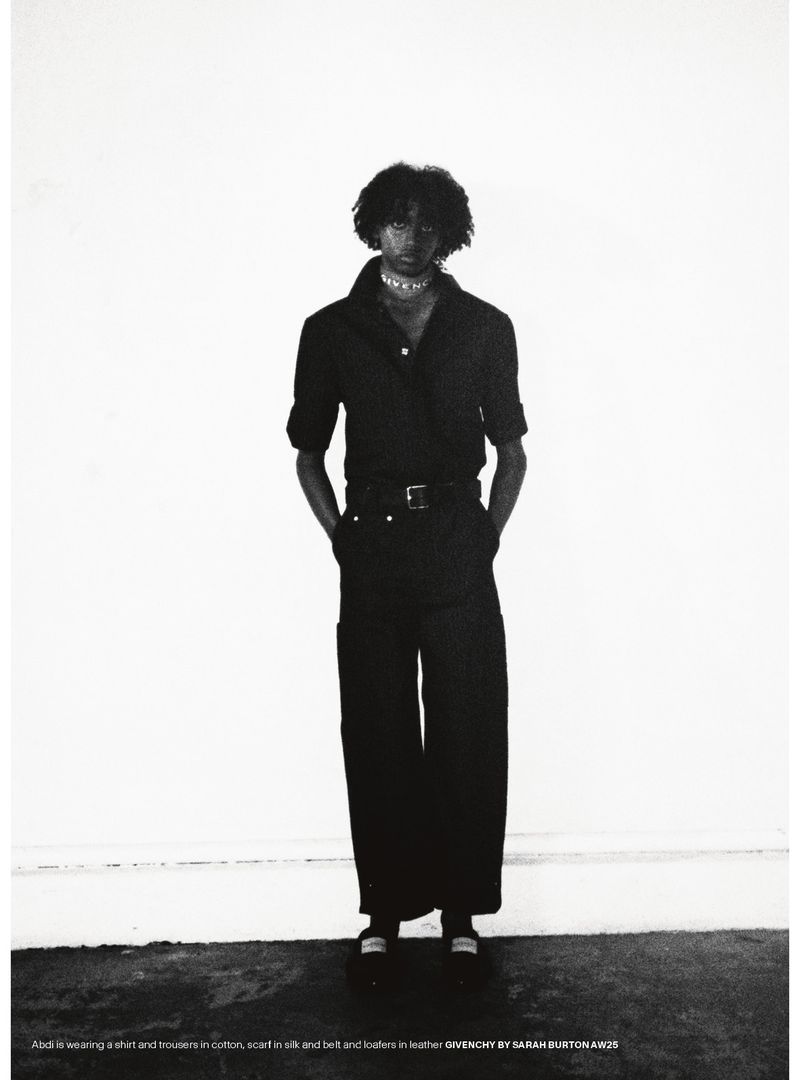 A slender adult is posing, wearing a black shirt, trousers, and jewelry against a stark white background in an indoor studio setting.