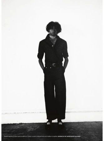 A slender adult is posing, wearing a black shirt, trousers, and jewelry against a stark white background in an indoor studio setting.