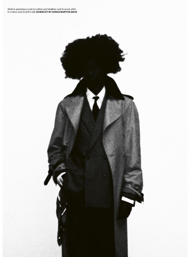 A person with an afro is wearing a coat, vest, tie, shirt, pants, gloves, and scarf, all in black and gray tones, in a studio shot.