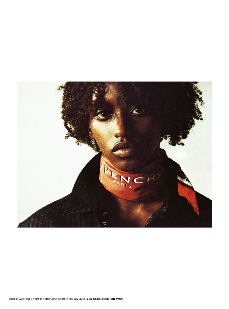 A young adult is posing indoors with a silk scarf wrapped around their neck and a black denim shirt in a fashion editorial shot.