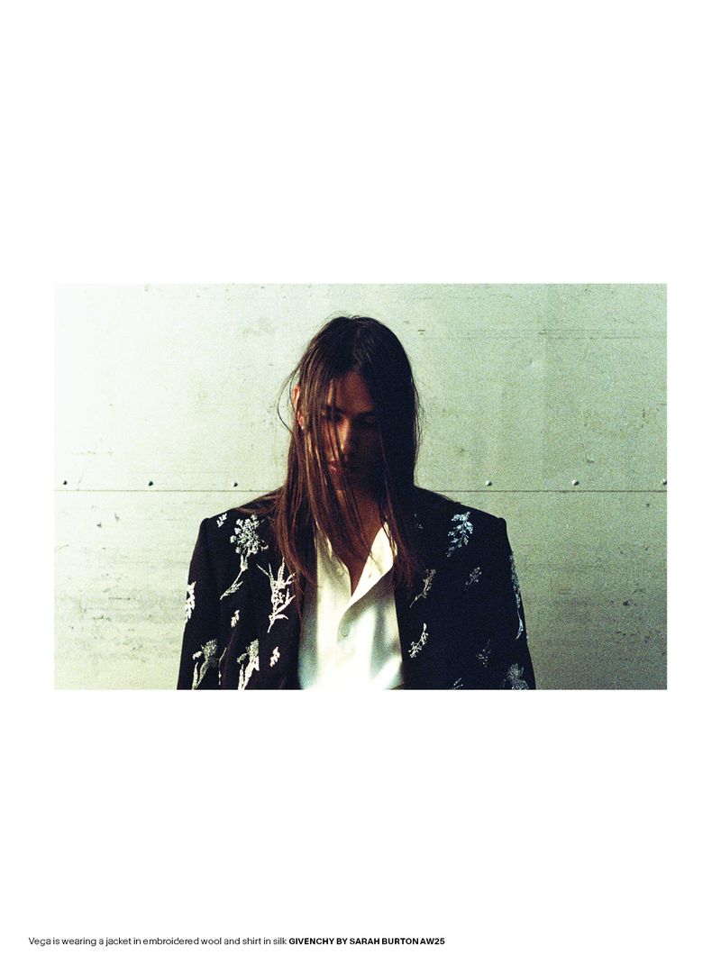 Adult male model with long brown hair wears an embroidered jacket and buttoned white shirt in a cool-toned indoor setting.