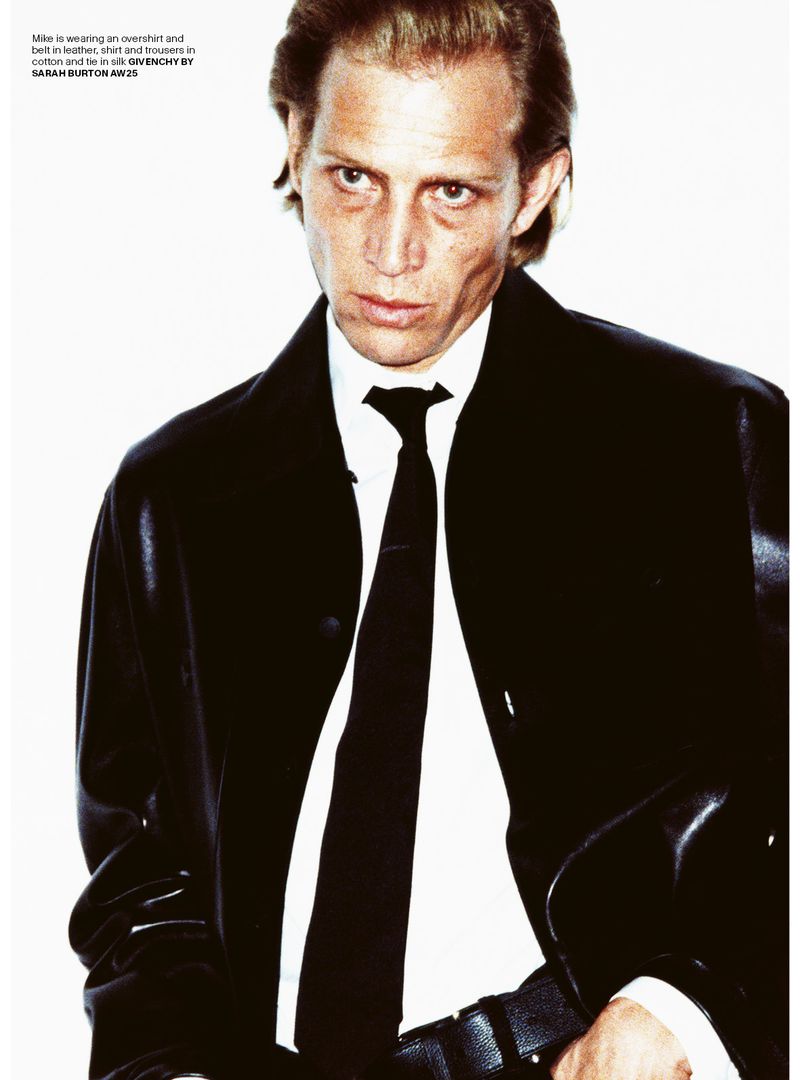 Man in black leather coat and tie poses in studio. Male fashion editorial shot against a white background, showcasing menswear.
