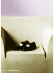 Motion-blurred pair of black leather loafers resting on a minimalist pale sculptural armchair against a soft purple wall.