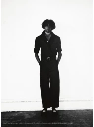 Black and white portrait of a model wearing a dark short-sleeved shirt and wide-leg trousers against a white wall.