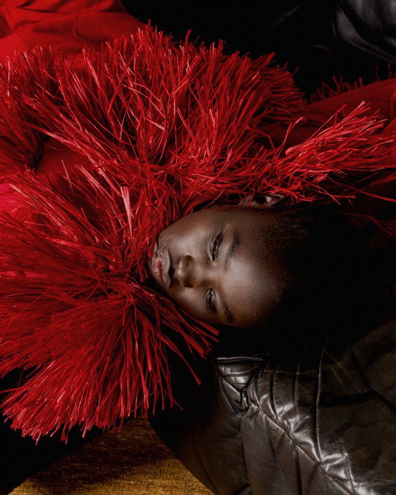 Portrait of a young African American child lying down, adorned with a vibrant red feathered garment against a contrasting black backdrop.