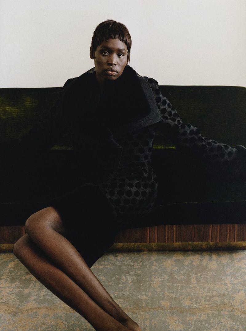 Elegant Black woman in a black polka dot jacket seated on green velvet couch against a white wall in a fashion editorial style.
