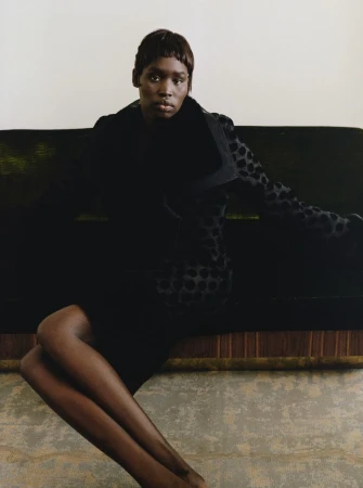Elegant Black woman in a black polka dot jacket seated on green velvet couch against a white wall in a fashion editorial style.