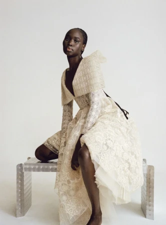 Elegant dark-skinned woman in a couture dress, seated on a metal bench in a light studio. Sophisticated fashion editorial.