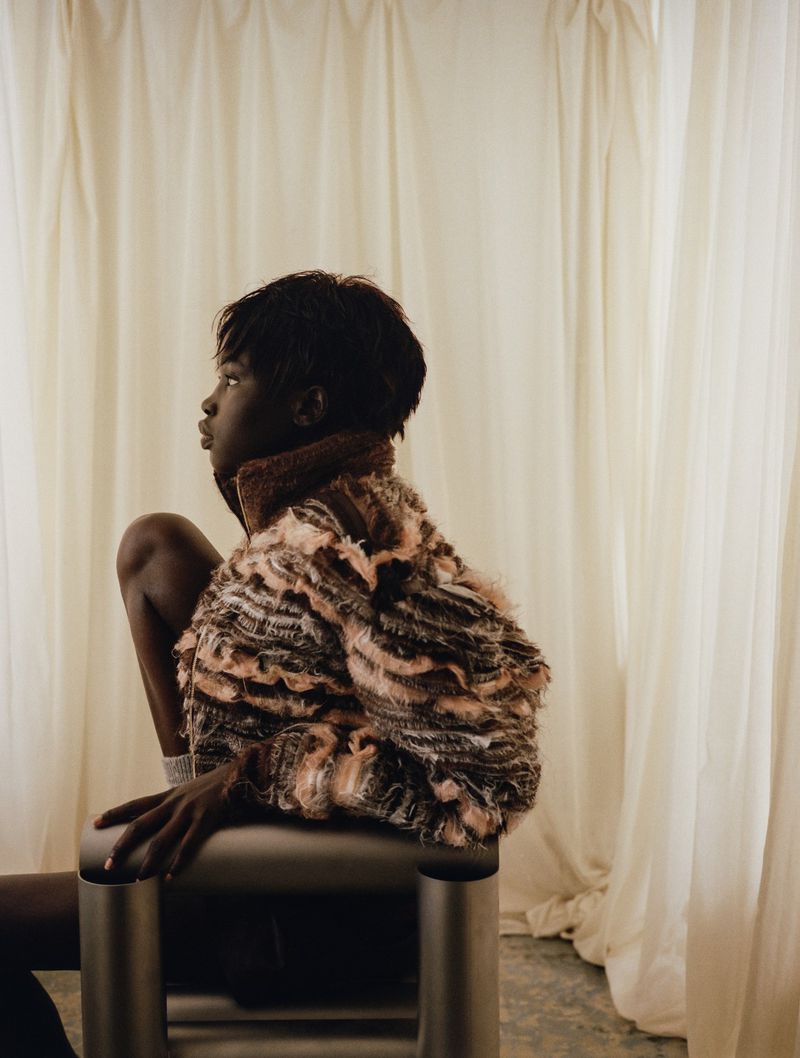 Child models a faux fur jacket while seated on a metal stool in front of draped white fabric in a studio setting.