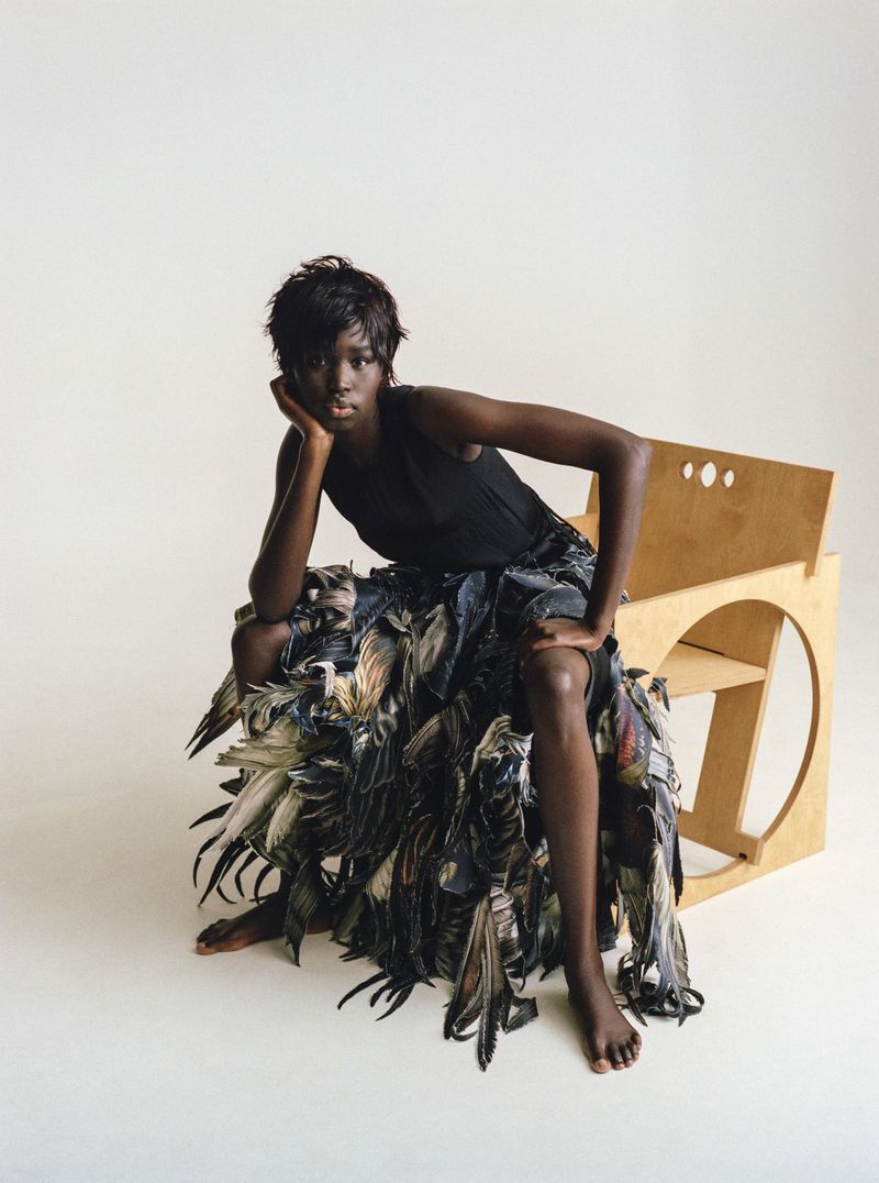 Fashionable adult woman with dark skin sits in an artful pose on a unique chair made of layered materials in a brightly lit photo studio.