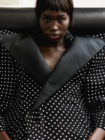 Black model with dark hair and bangs wearing a black and white polka dot garment against a dark background