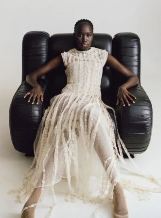 Model wearing cream lace dress seated in a large black leather armchair against a plain white background