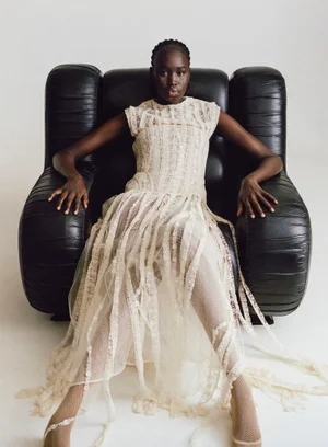 Model wearing cream lace dress seated in a large black leather armchair against a plain white background