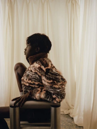 Profile of a black woman seated on a metallic chair wearing a textured patterned jacket against cream curtains