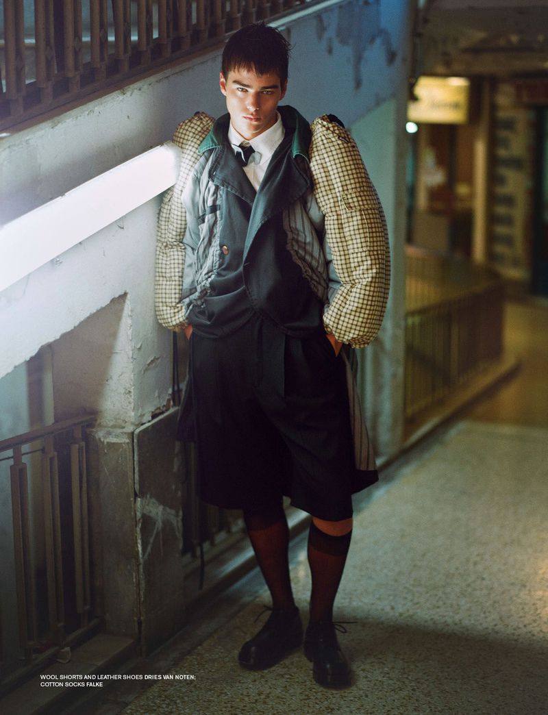 Fashionable young man in a stylish dark jacket and shorts posing in an urban hallway with a serious facial expression, editorial fashion.