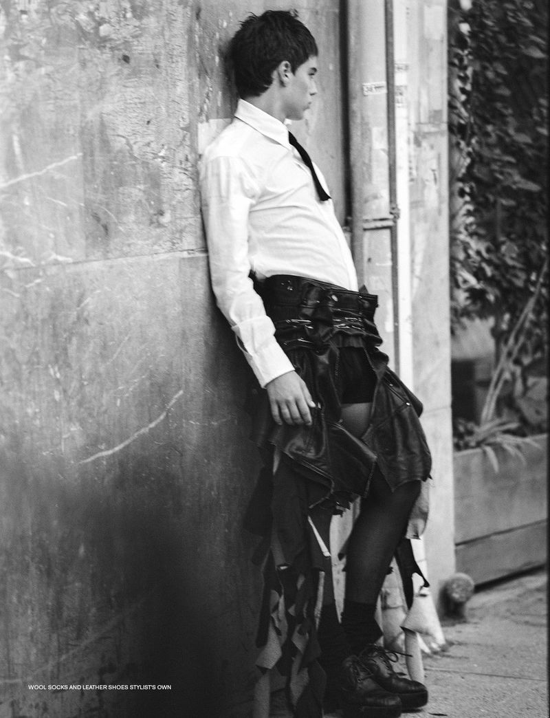 Black and white portrait of an adult male in white shirt, black tie, leather skirt, black stockings and shoes leaning against a textured wall.