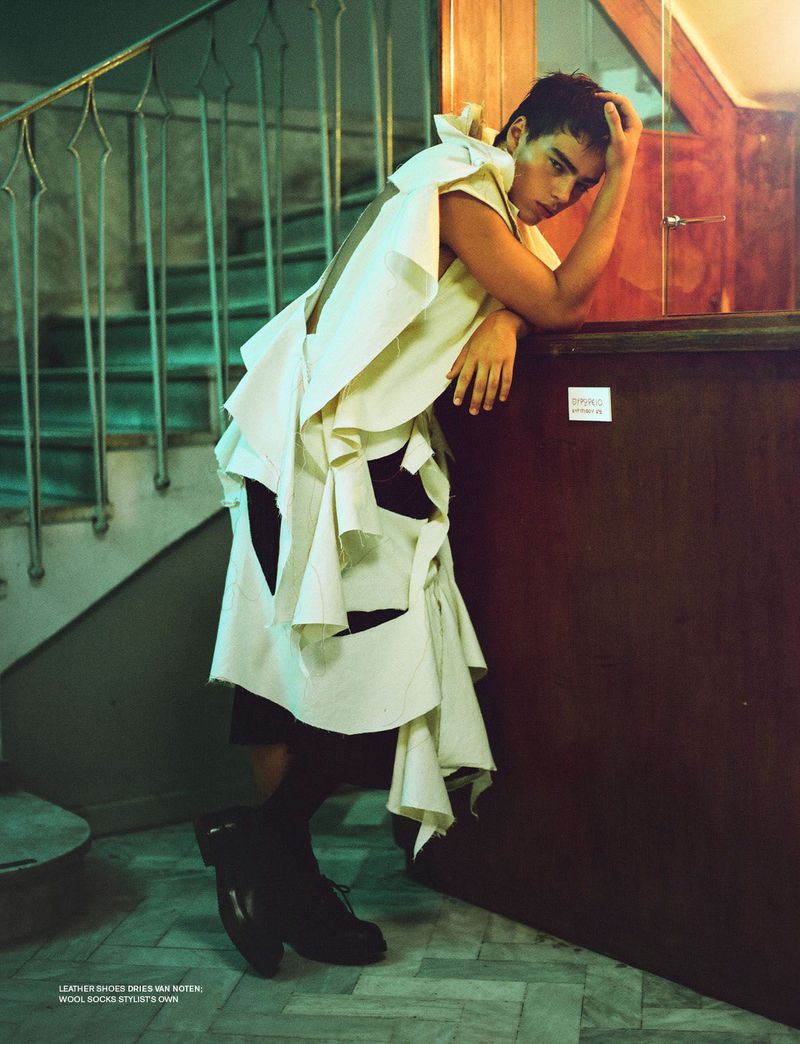 Fashion photo of a young adult with hand on head, posing in an ornate ivory top and skirt, near a dark wood paneled wall and stairway.