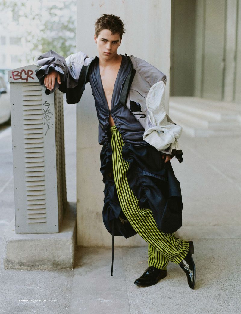Male fashion model posing in stylish avant-garde clothing in an urban setting, leaning against a gray utility box on a city street.